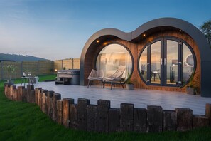 Una casa moderna con amplios ventanales circulares, una terraza de madera y mobiliario exterior.