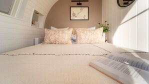 Un dormitorio con cama, almohadas, una mesita de noche con una planta y un cuadro en la pared.