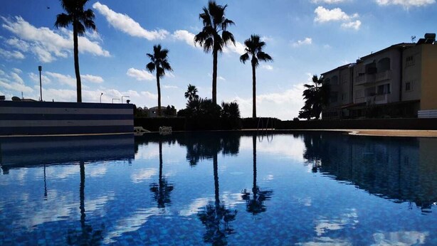 A swimming pool with clear water reflecting palm trees and a building in the background. - Escape and Relax Apartment (Mandria)