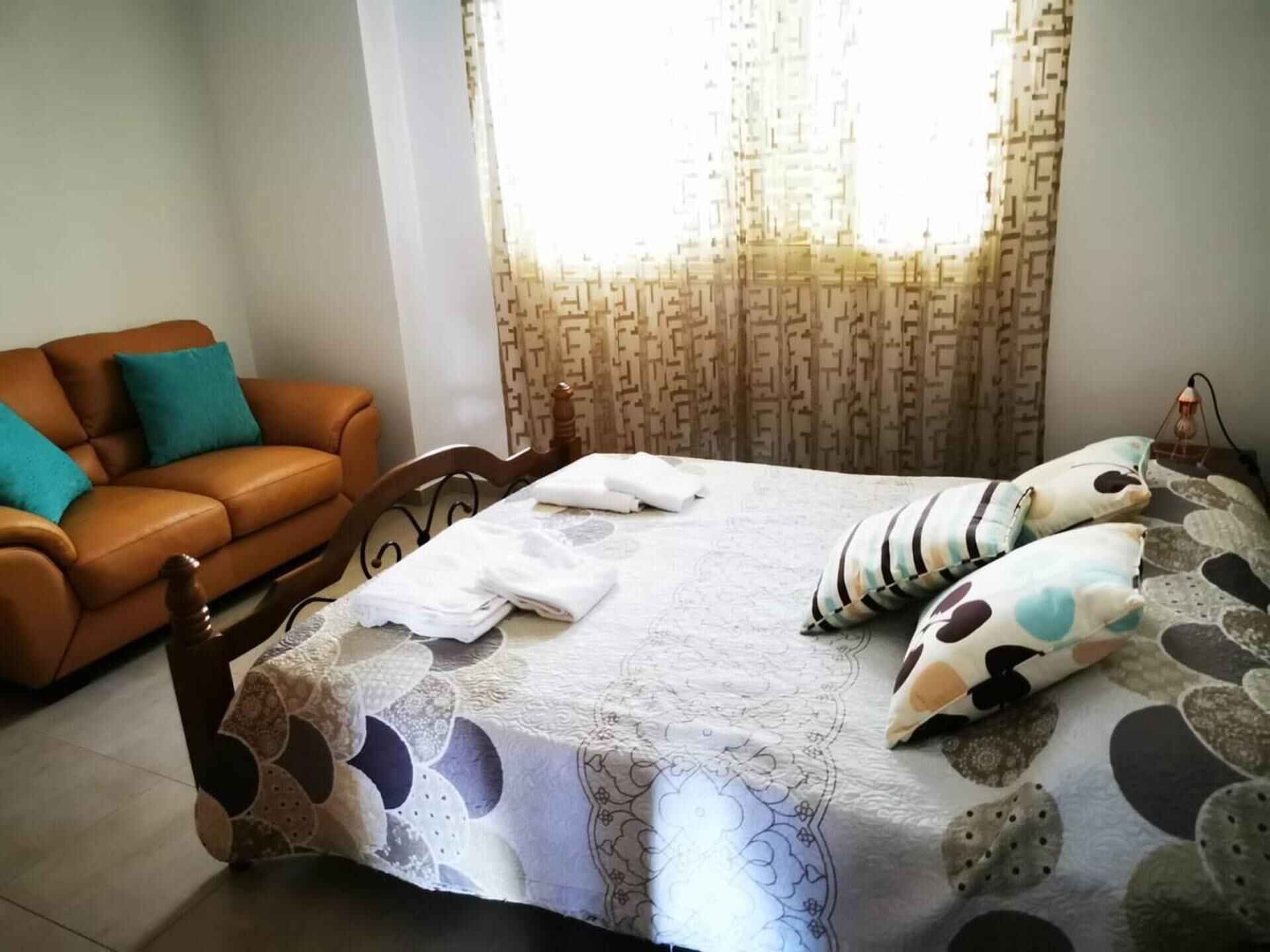 A bedroom with a bed, a wooden headboard, a brown leather sofa, and a patterned curtain.