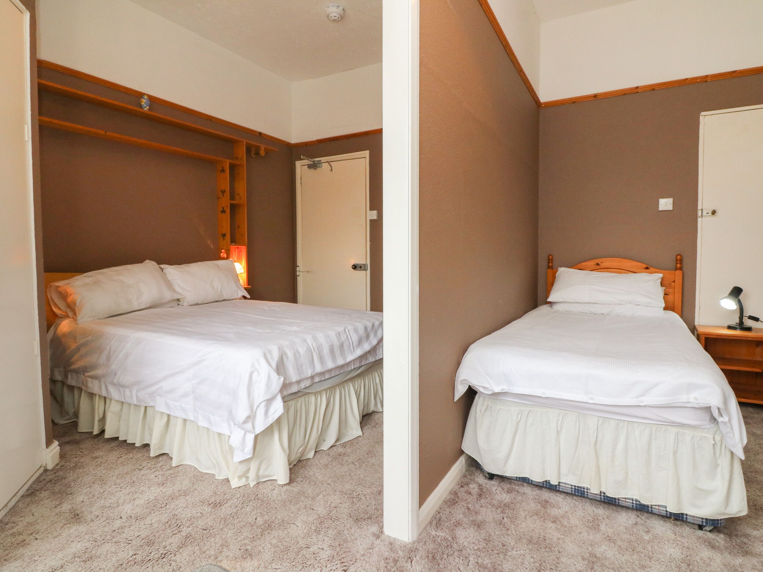 Une chambre avec deux lits, une tête de lit en bois, une table de chevet avec une lampe et une porte donnant sur une autre pièce.
