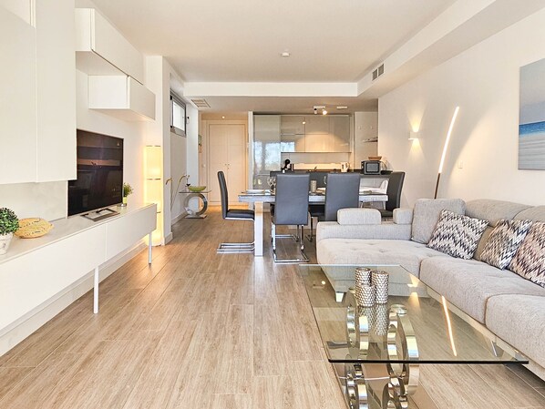 A modern living room with a glass coffee table, a grey sofa, and a flat-screen TV mounted on the wall.
