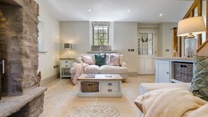 A cozy living room with a stone fireplace, a white sofa with decorative pillows, a wooden coffee table, and a view of a window with a wreath. - Brontë Cottage  -  3 Bedroom Riverside Cottage. Sedbergh 4 Miles. (Garsdale)
