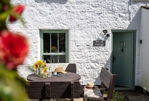 A rustic cottage with a stone exterior, a green door, and a table with sunflowers and a hat.