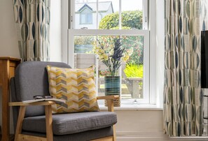 A room with a window, a chair with a cushion, a small table with glasses and a book, and a vase with flowers.