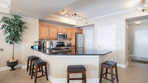 A modern kitchen with wooden cabinets, a central island with stools, a potted plant, and a stainless steel refrigerator.