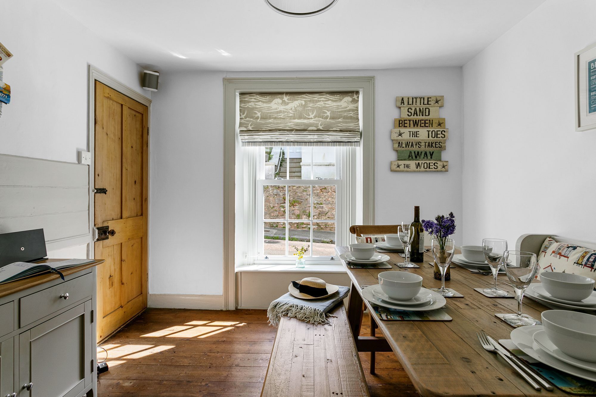 Une cuisine avec une table en bois dressée pour le repas, une fenêtre offrant une belle vue et un mur orné d’un panneau décoratif.