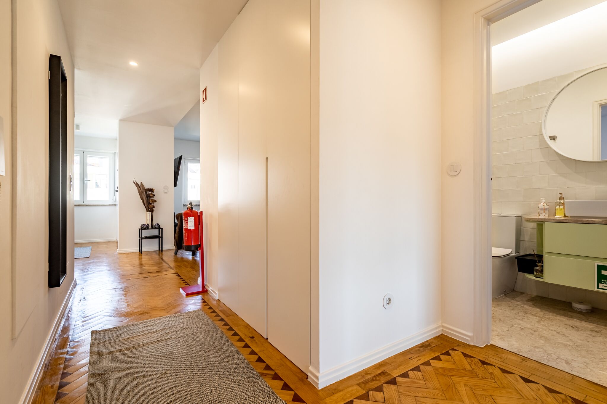 Un couloir doté d’un plancher en bois, de murs blancs et d’une salle de bain verte avec un lavabo et un miroir blancs.