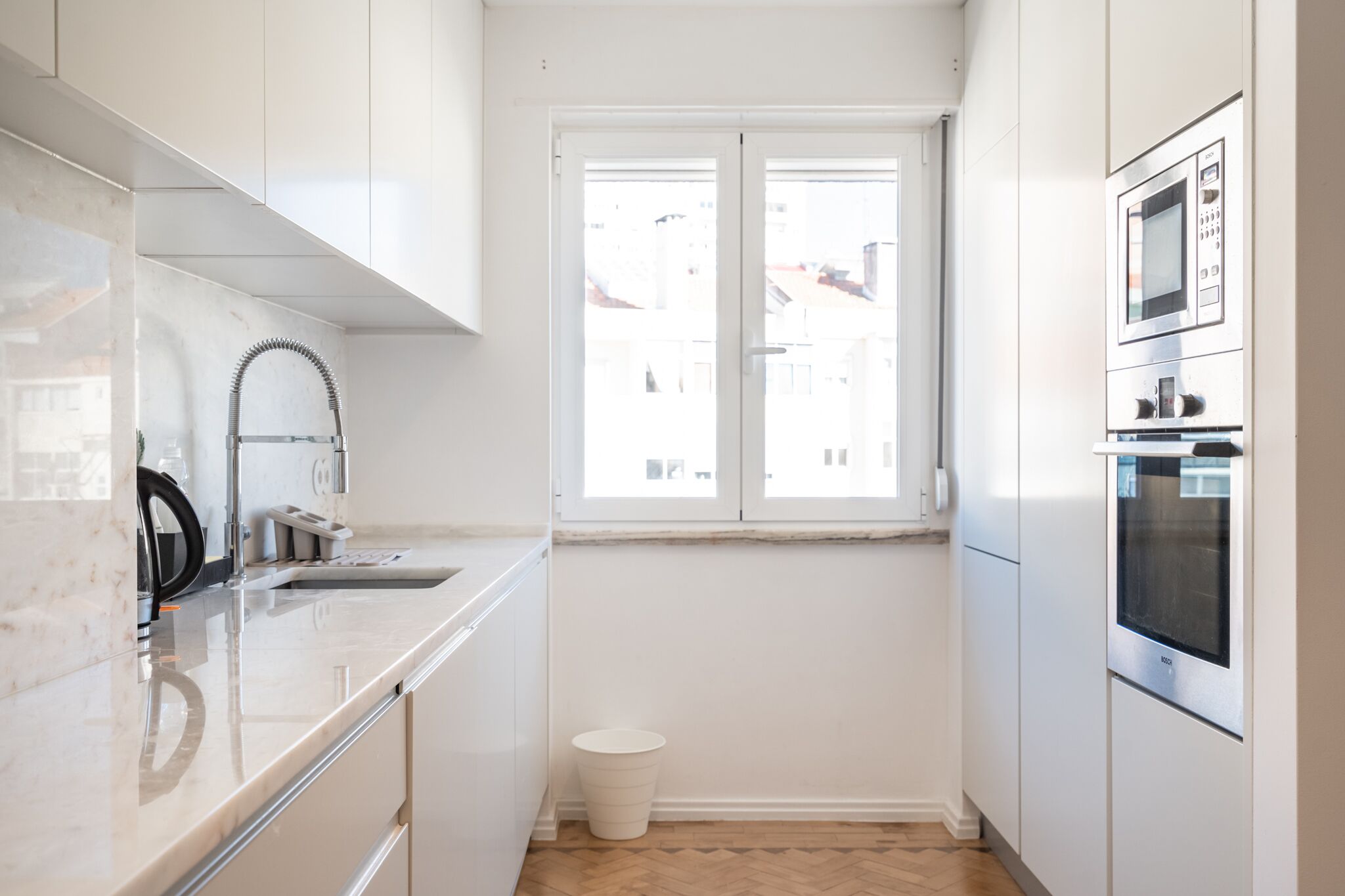 A modern kitchen with white cabinetry, a stainless steel sink, a built-in microwave, and a window with a view of buildings outside.