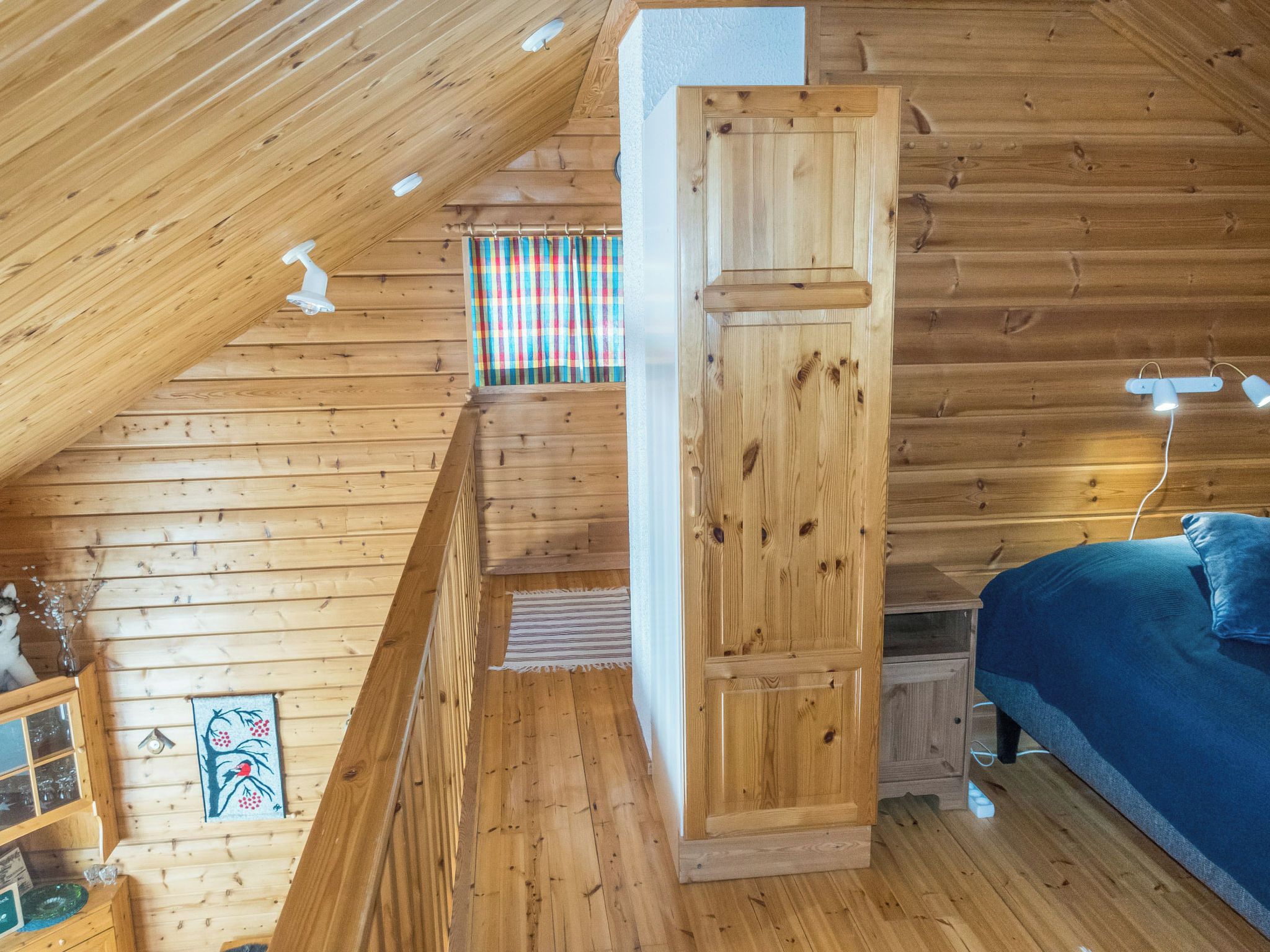 Interior de una cabaña de madera con techo inclinado, una cama con almohadas, una ventana con cortinas y una pequeña mesa con vasos.