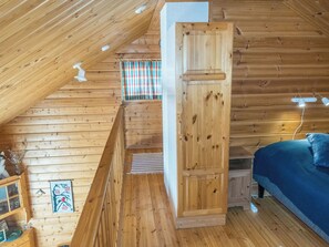 Ein Holzblockhaus-Interieur mit schrägem Dach, einem Bett mit Kissen, einem Fenster mit Vorhängen und einem kleinen Tisch mit Gläsern.