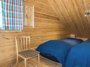 Ein Holzblockhaus-Innenraum mit einem Einzelbett, einem Sessel und einem Fenster mit einem Karomuster-Vorhang.