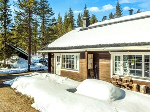 A wooden cabin with a snow-covered roof, large windows, and a wooden deck with outdoor furniture.