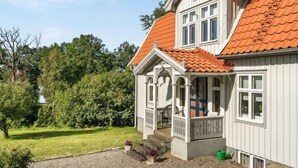 Une maison à deux étages avec un toit recouvert de tuiles rouges, une véranda dotée d’une balustrade et un petit jardin agrémenté d’une allée en gravier.