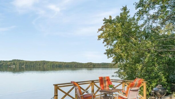 Une terrasse en bois avec mobilier d’extérieur, un barbecue et une vue sur un lac paisible et une forêt.