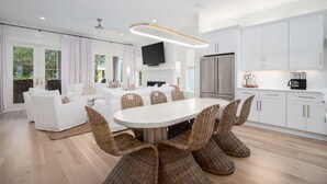 A modern kitchen with white cabinets, a stainless steel refrigerator, and a dining area with wicker chairs and a white table. - Over the Moon, 30A Home, Golf Cart, Elevator, Pool (Santa Rosa Beach)