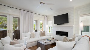 A modern living room with white furniture, a flat-screen TV mounted above a fireplace, and large windows with curtains. - Over the Moon, 30A Home, Golf Cart, Elevator, Pool (Santa Rosa Beach)