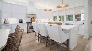A modern kitchen with white cabinetry, a central island, and a dining area with wicker chairs. - Over the Moon, 30A Home, Golf Cart, Elevator, Pool (Santa Rosa Beach)