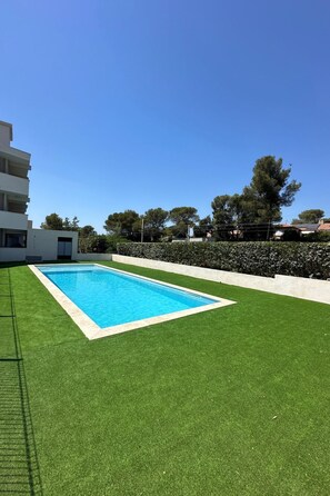 Ein rechteckiger Swimmingpool mit weißem Rand, umgeben von grünem Kunstrasen, vor blauem Himmel.