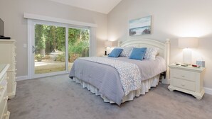 Een slaapkamer met een grote schuifpui naar een tuin, een wit bed met beddengoed met een patroon, een nachtkastje met een lamp en een plafondventilator.