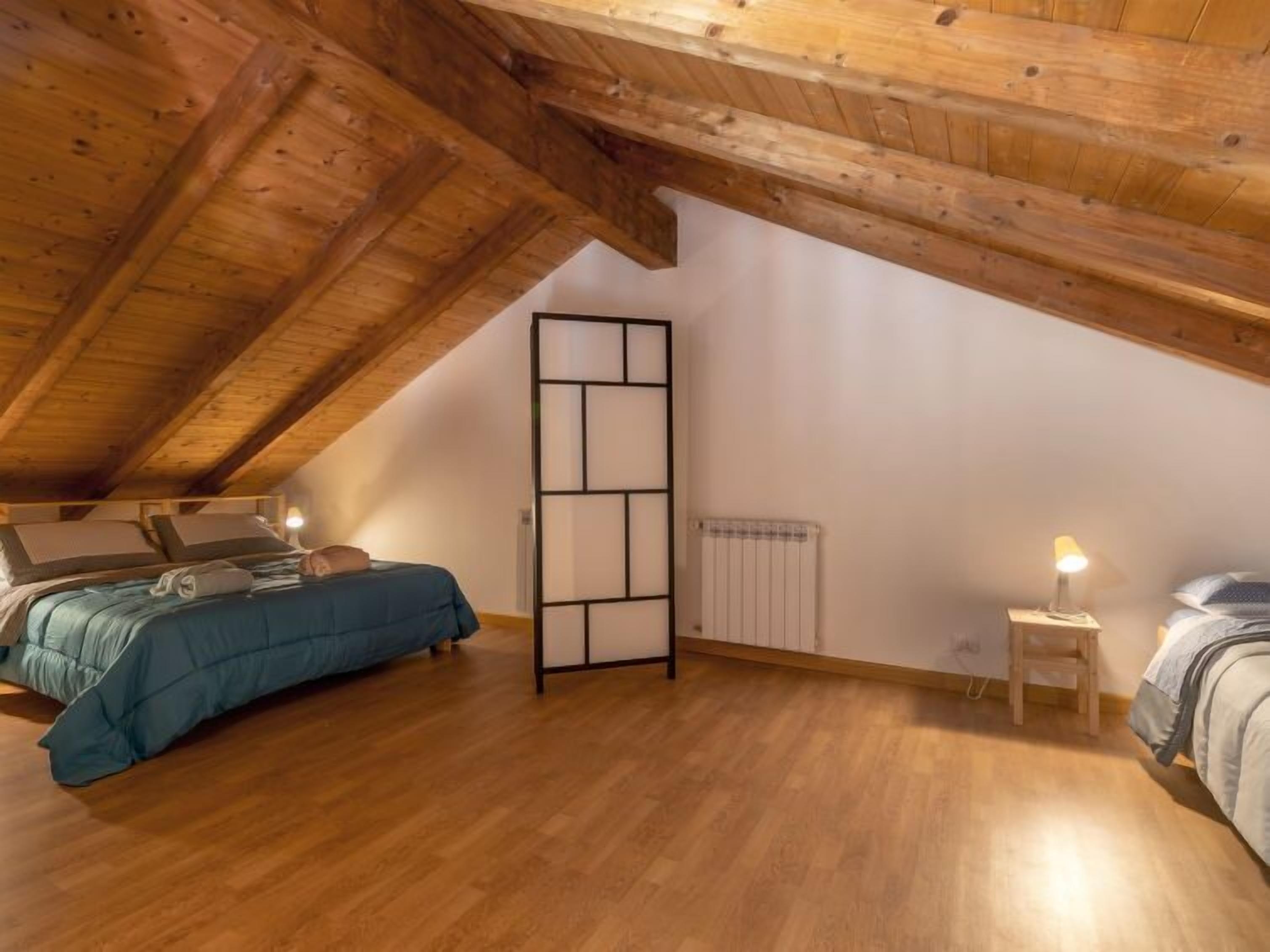 A bedroom with a sloped ceiling, wooden beams, a bed with a blue comforter, a radiator, and a bedside table with a lamp.
