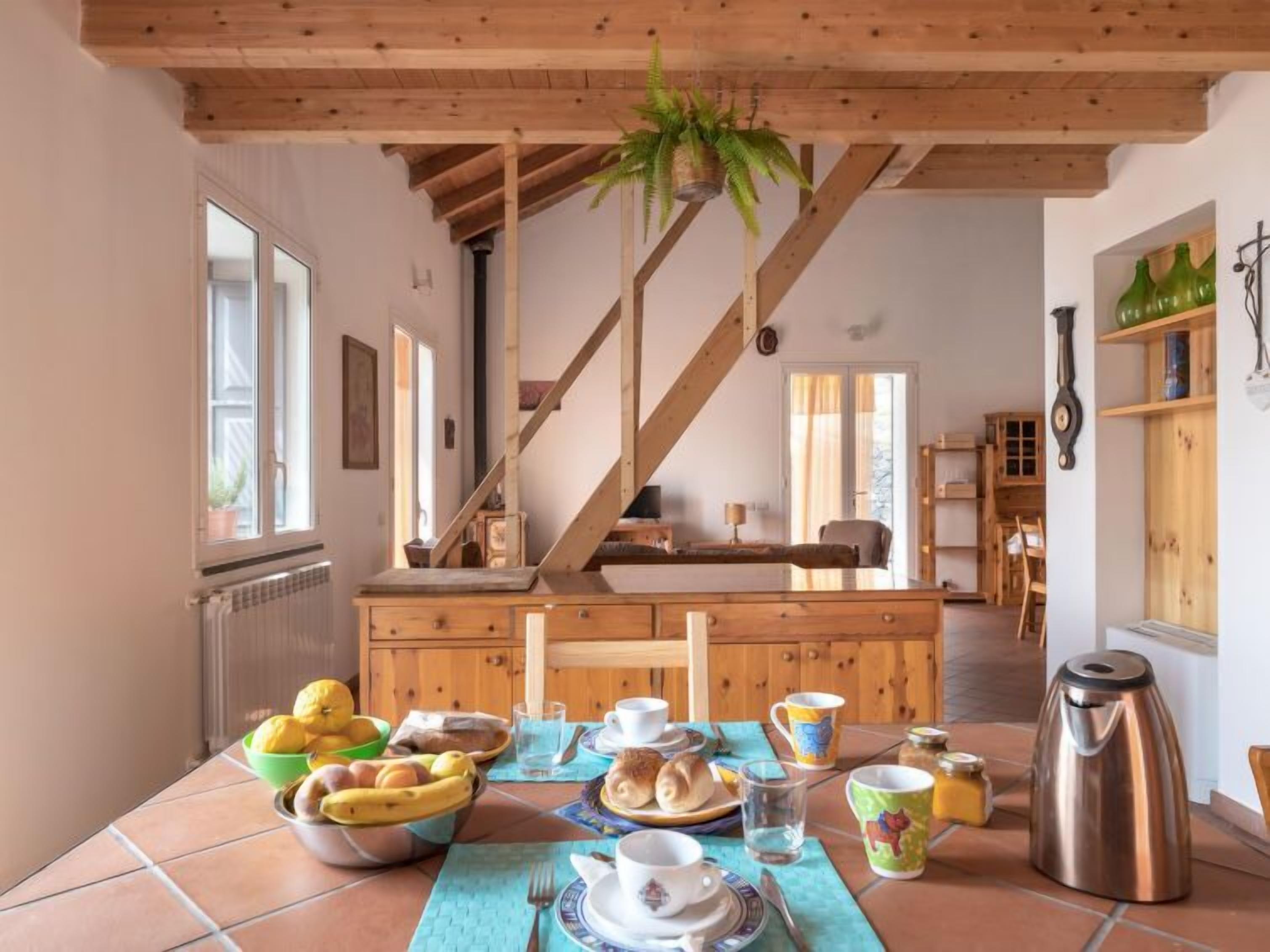 A dining area with a wooden table set for breakfast, a staircase, and a view of the kitchen.