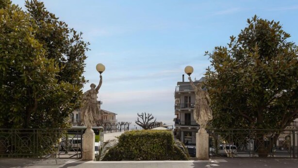 A paved walkway with a statue on each side, flanked by trees and a building in the background.