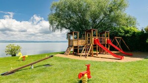 Un parque infantil con una estructura de madera, toboganes y columpios, frente a un lago y árboles.