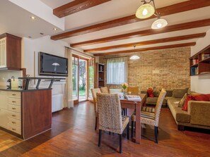 A spacious living area with a wooden ceiling, a dining table with chairs, a sofa, and a television mounted on the wall.