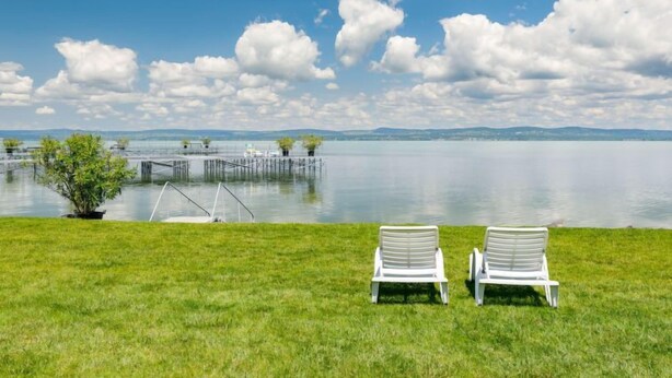 Dos sillas de lounge blancas mirando un lago tranquilo con un muelle y pequeños árboles al fondo.