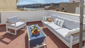 Eine Dachterrasse mit einem weißen Sofa, zwei weißen Stühlen, einem Tisch mit Obstschale und Blick auf eine Küstenstadt.