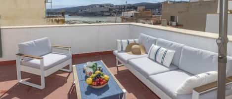 A rooftop terrace with a white sofa, two white chairs, a table with a fruit bowl, and a view of a coastal town.