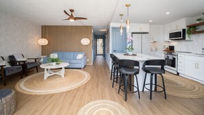 A modern living room with a sofa, chairs, a dining area with bar stools, a kitchen with white cabinets, and a ceiling fan. - Maui Parkshore 204 2BR across Kamaole III Beach w/ ocean views, summer deal (Kihei)