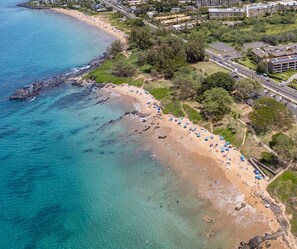 Une vue aérienne d’une plage aux eaux turquoise et limpides, avec un rivage sablonneux et une rangée de parasols de plage.