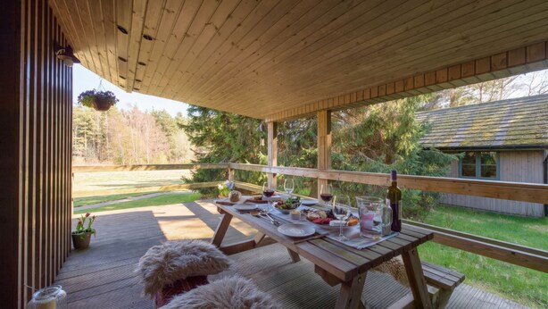 A wooden deck with a table set for dining, overlooking a grassy area and trees. - Cosy, comfortable lodge sleeps 6 guests  in 3 bedrooms. Pet friendly! (Banchory)