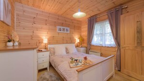 A cozy wooden cabin room with a bed, bedside tables, a window with curtains, and a hanging light fixture. - Cosy, comfortable lodge sleeps 6 guests  in 3 bedrooms. Pet friendly! (Banchory)
