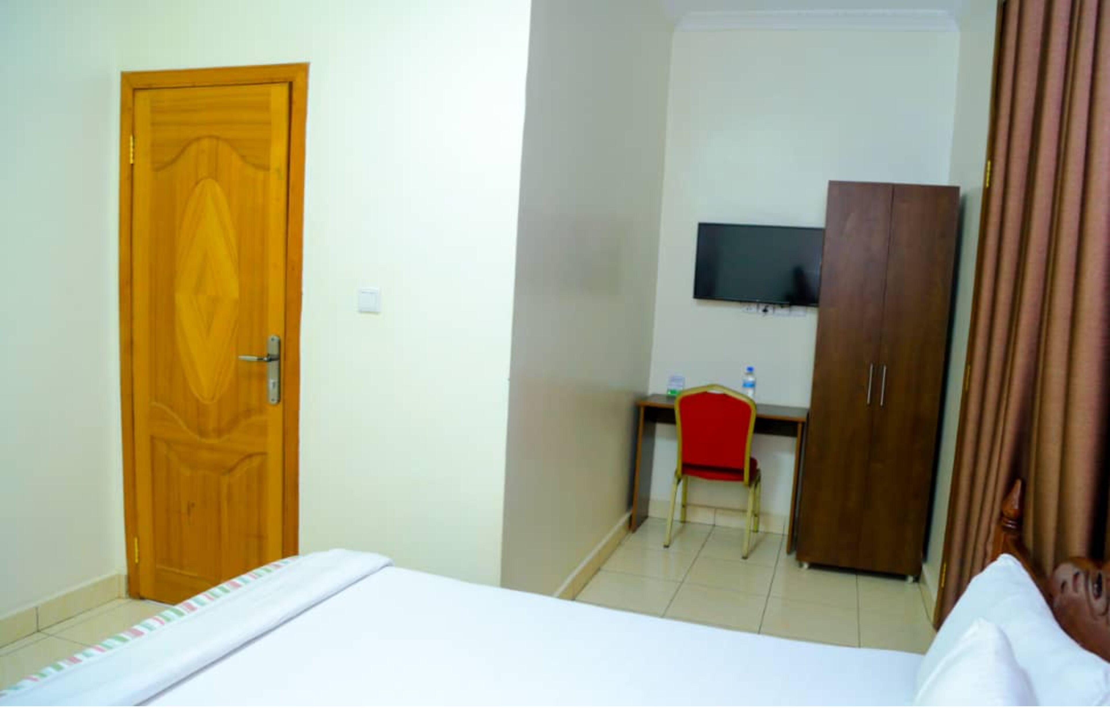 A hotel room with a wooden door, a bed with a white bedspread, a desk with a chair, a television on the wall, and a wardrobe.