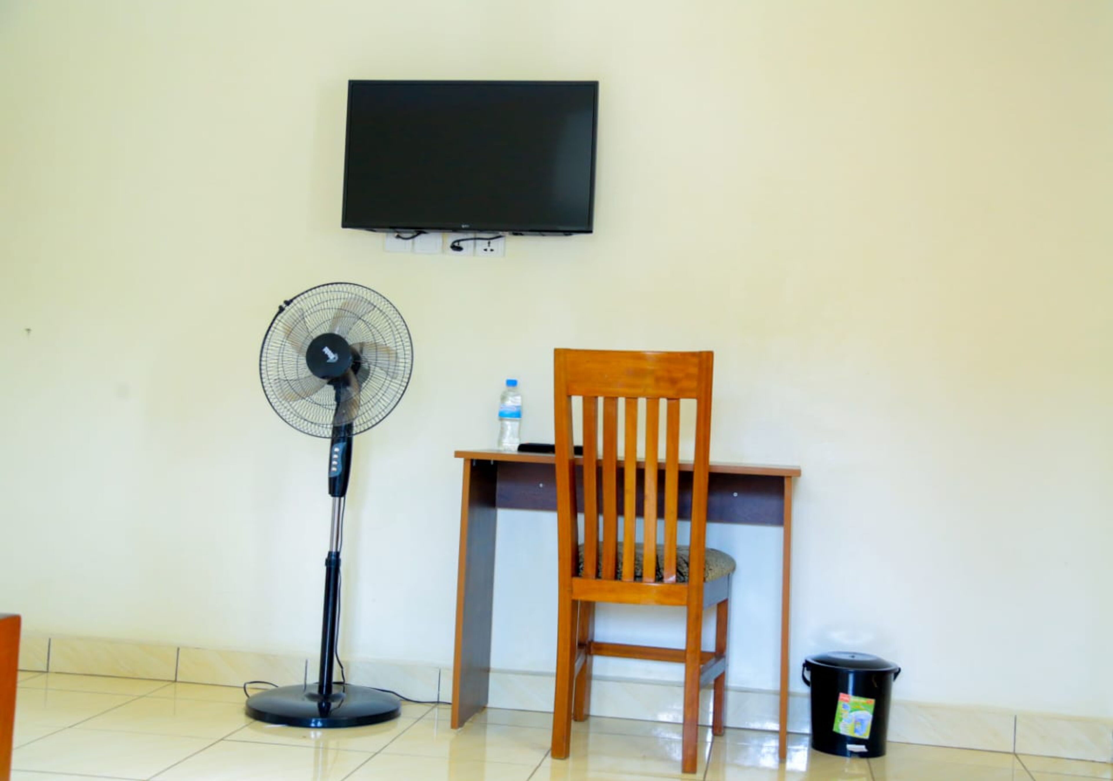 Una habitación con un escritorio de madera, una silla, un ventilador de pie, un televisor y un cubo de basura.