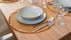 Tavola imbandita con piatti, bicchieri e posate, un sottotavolo in fibra naturale e un piccolo vaso con una pianta al centro.
