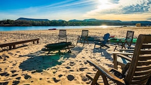 A beach with several wooden chairs and a bench, a boat, and a clear view of the water and distant hills. - Beachfront Villa | Concierge Services (Kingsland)