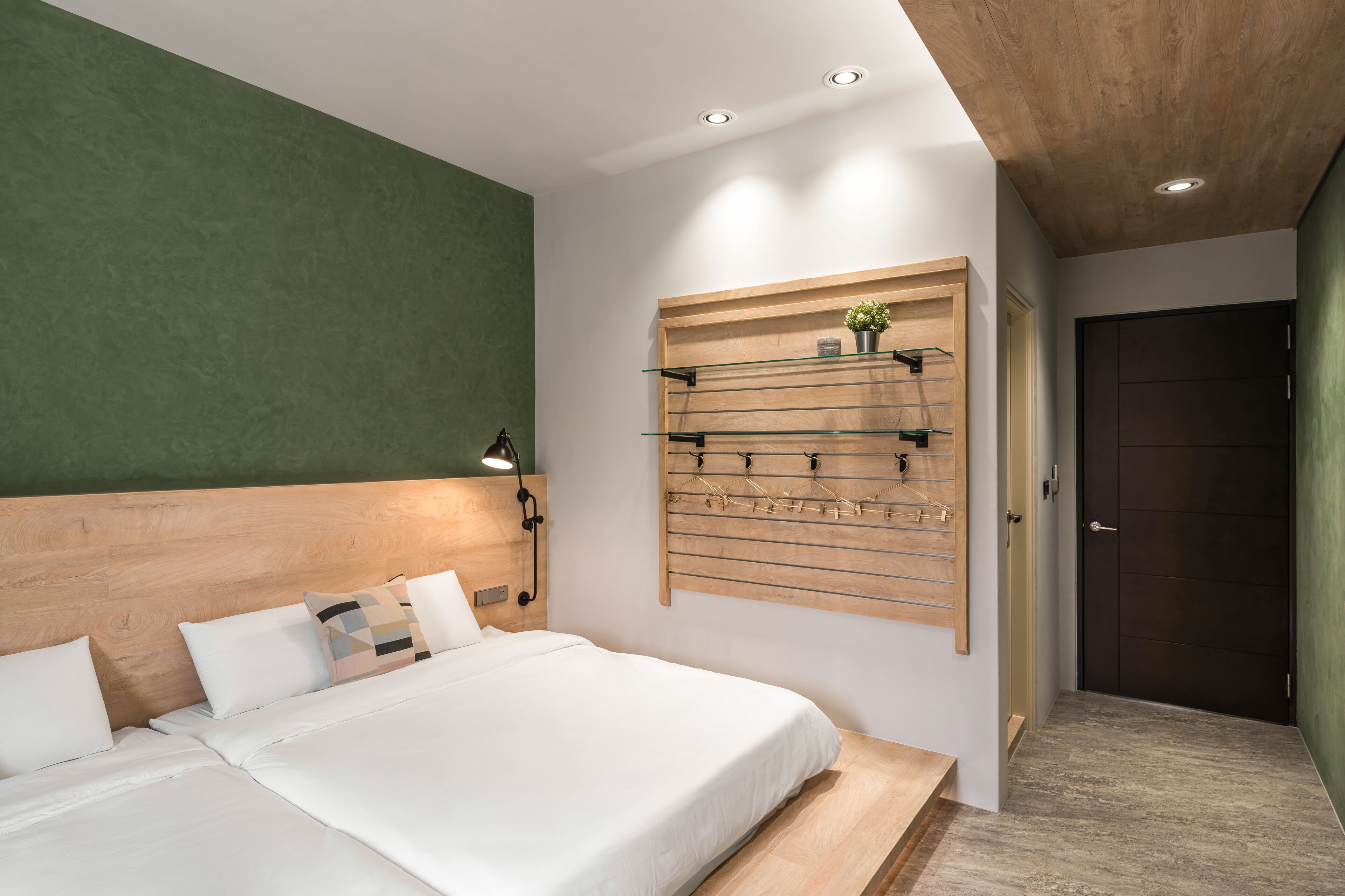 A modern hotel room with a large bed, a wooden headboard, a wall-mounted shelf with hooks, and a dark door.