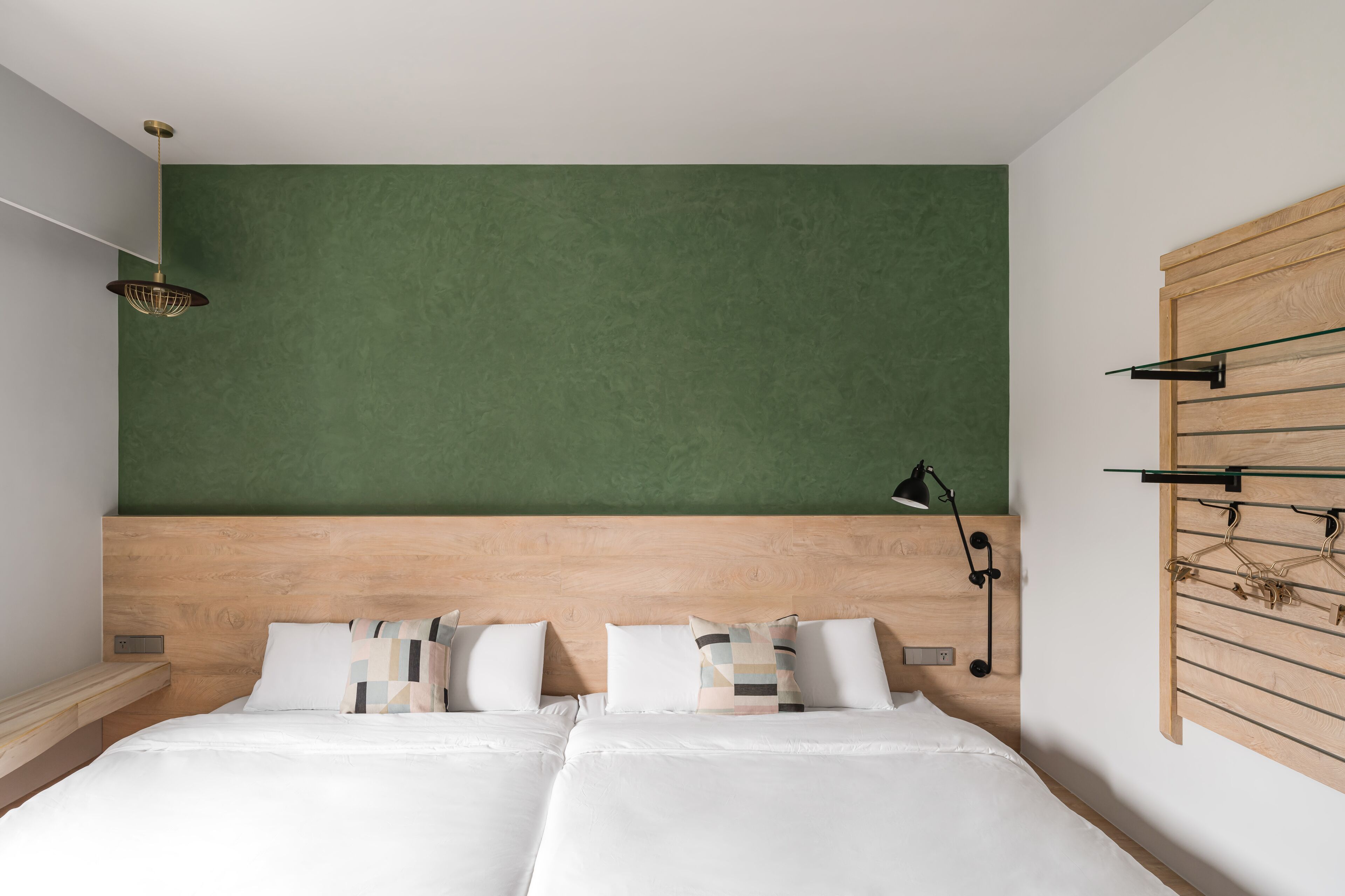 Habitación de hotel moderna con cabecera de madera, dos camas con ropa blanca, una pared de acento verde y un perchero con perchas.