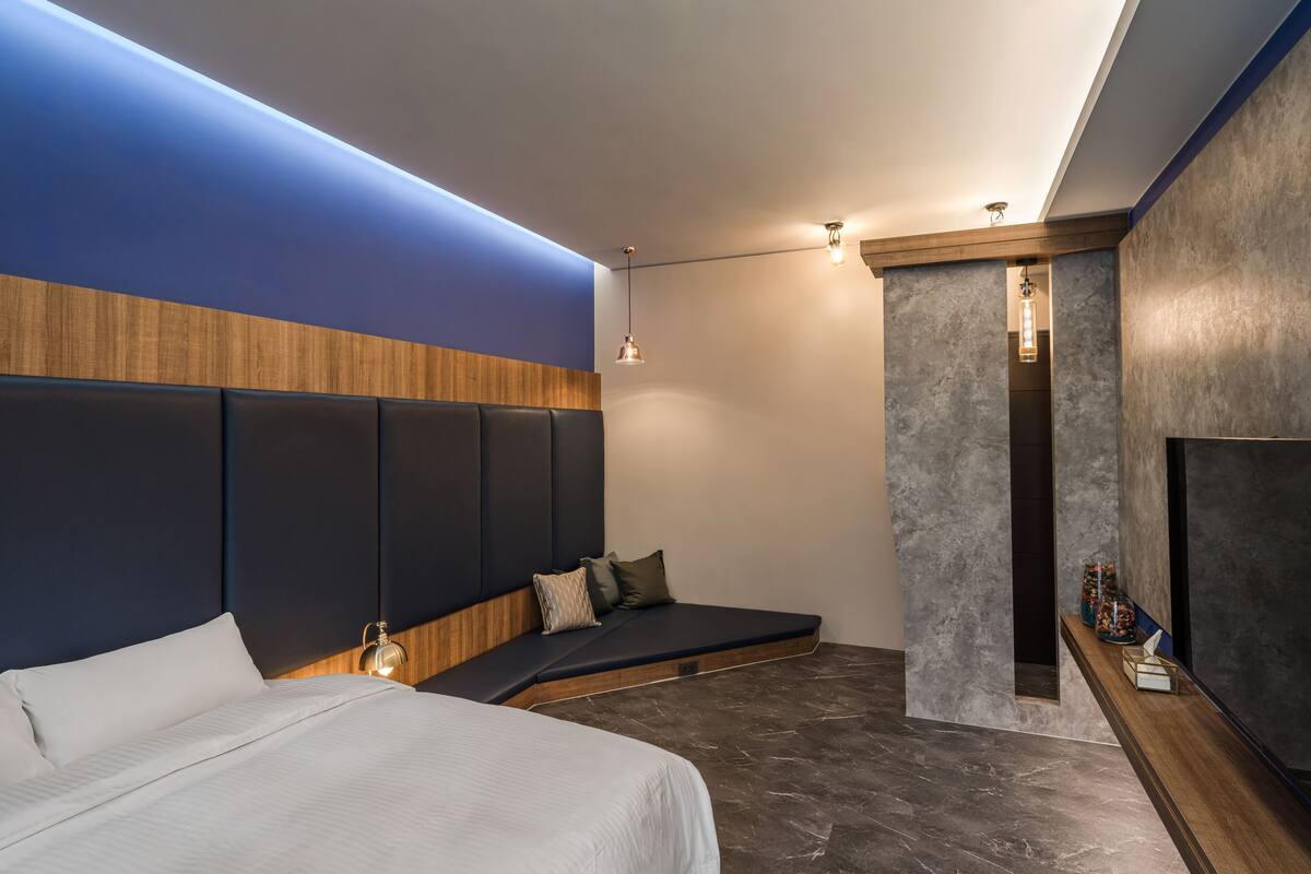 A modern hotel room with a large bed, a built-in bench, a TV, and a small shelf with items.