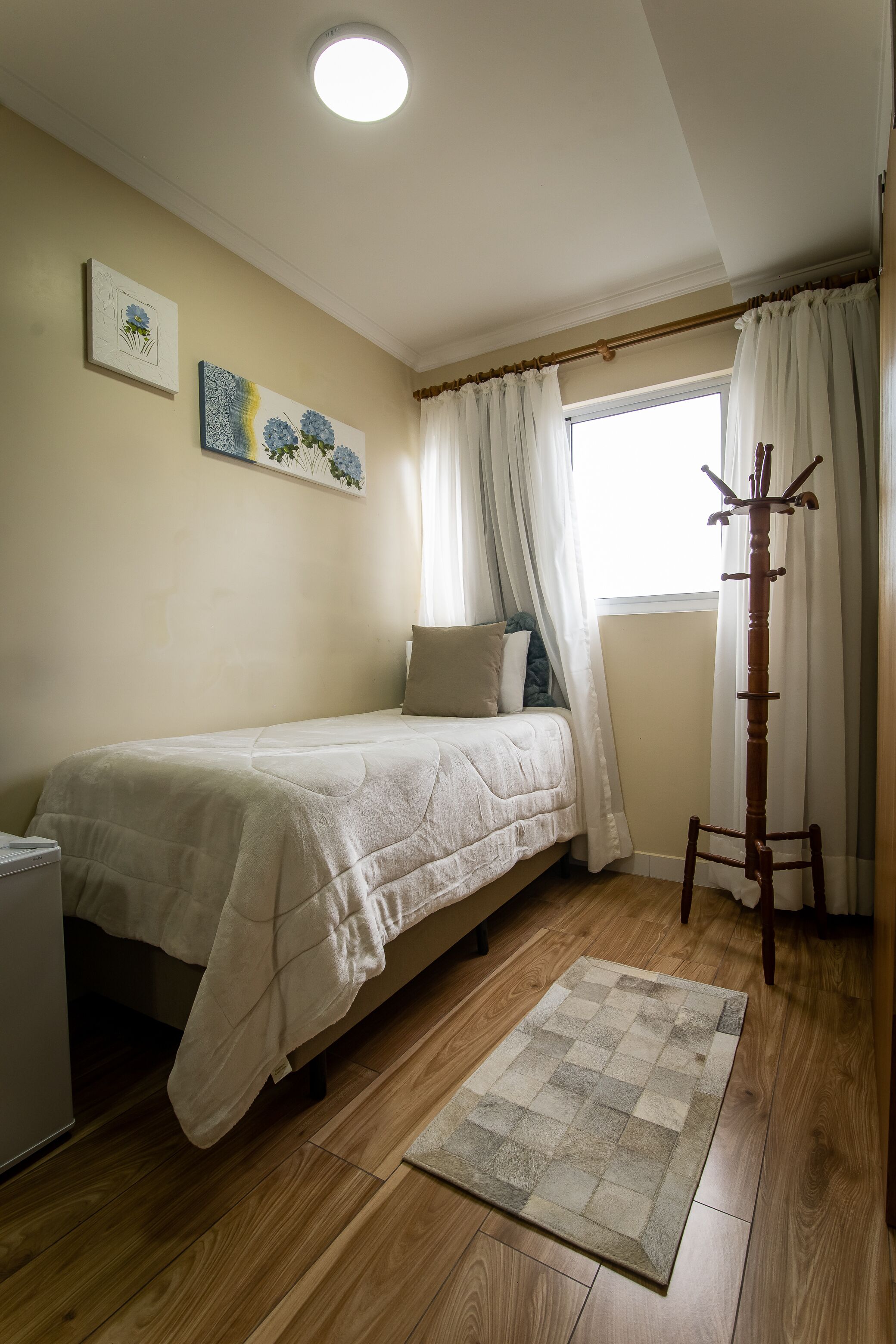 A bedroom with a bed, a wooden floor, a window with curtains, and a coat rack.