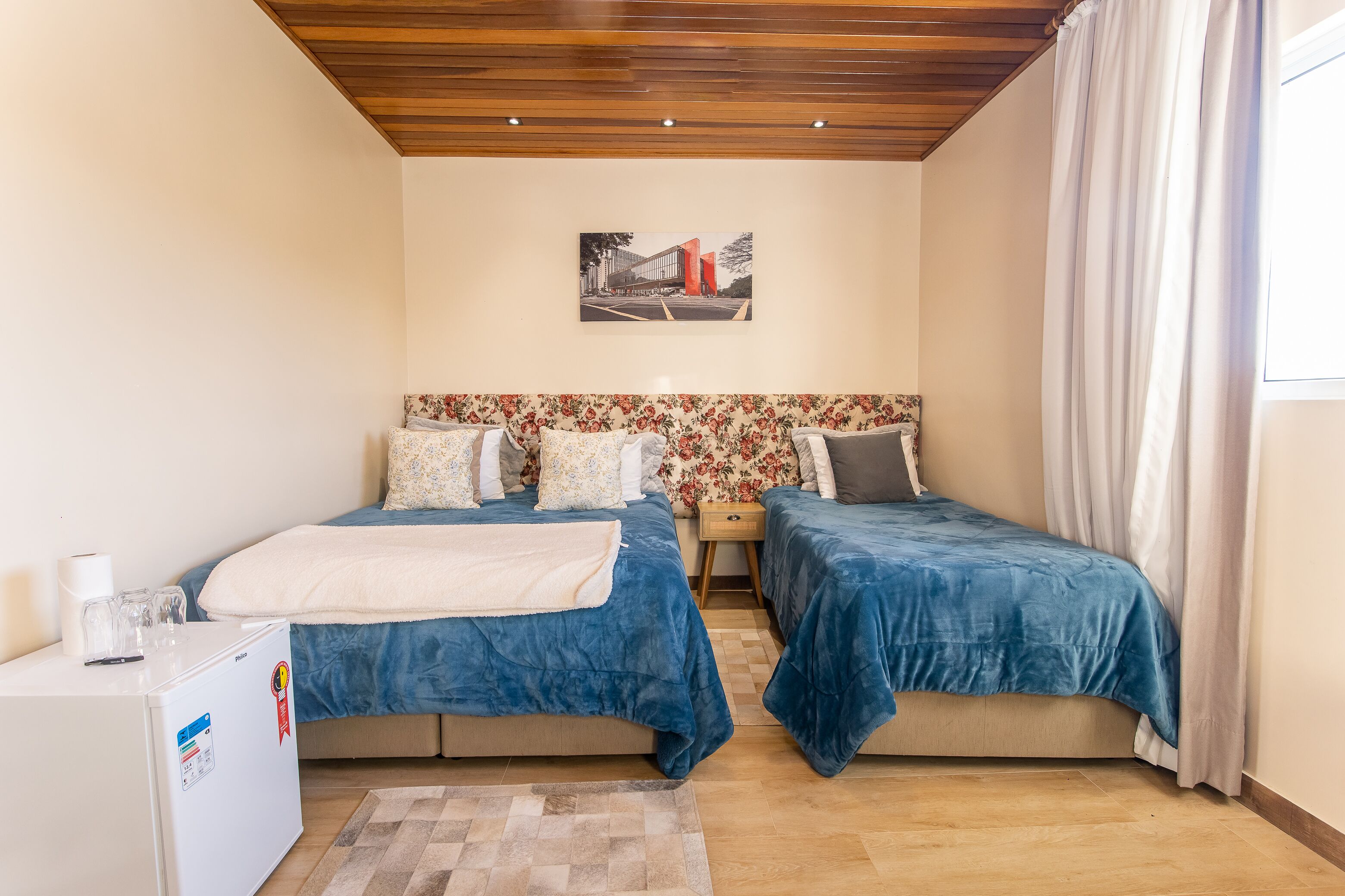 A bedroom with two beds, a small white desk, a wooden ceiling, a floral headboard, and a picture on the wall.
