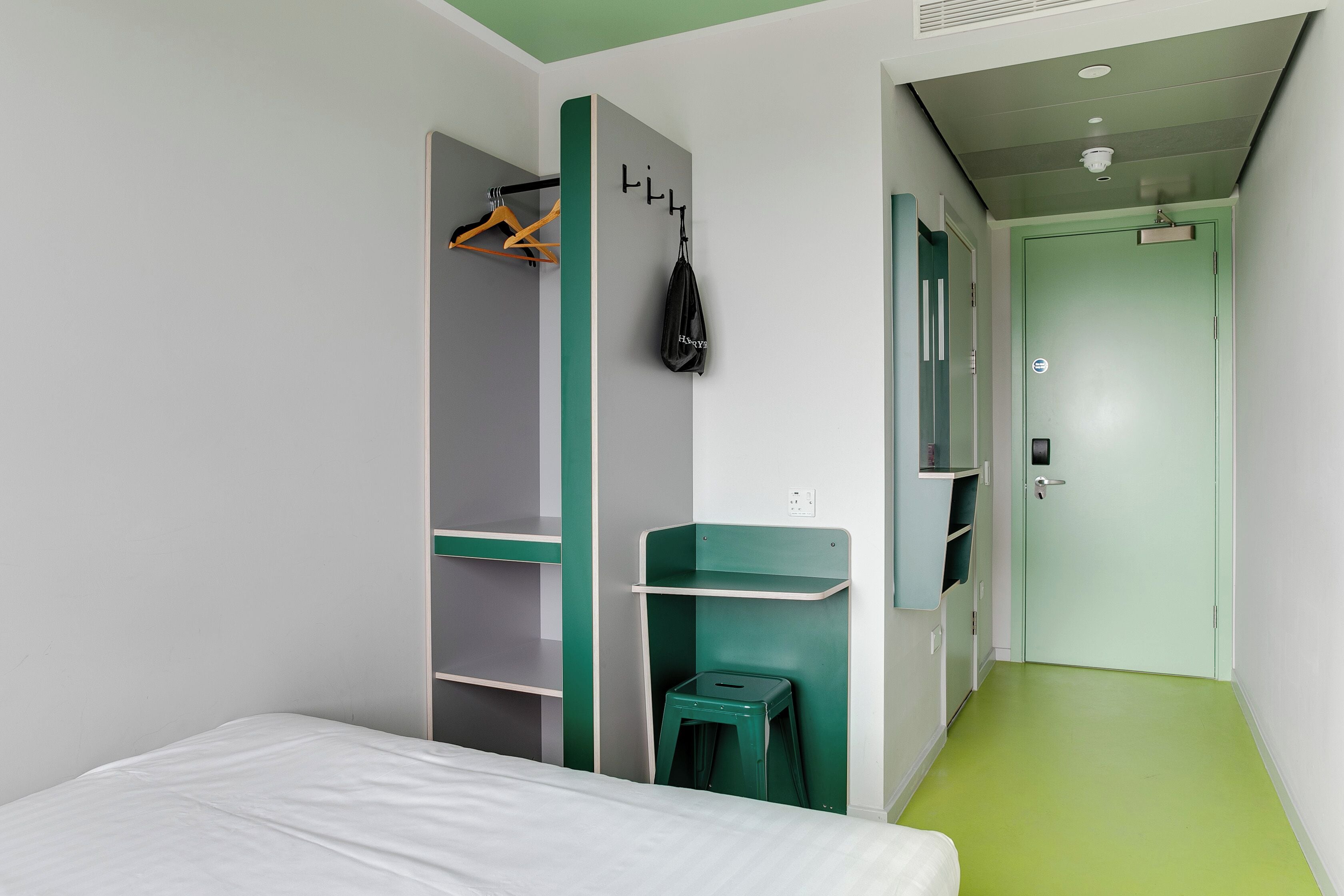 A hotel room with a bed, a green stool, a coat rack with hangers, and a green wall unit with shelves.