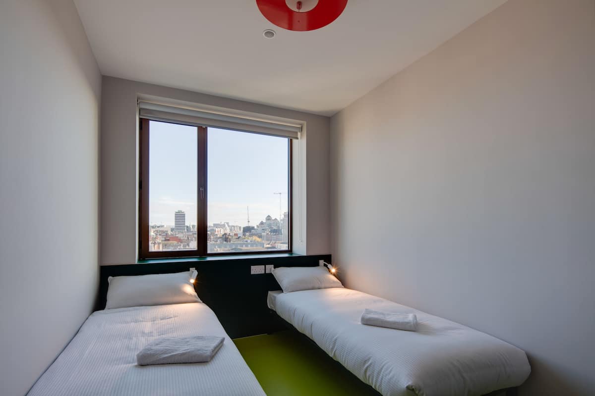 A modern hotel room with two single beds, a large window with a city view, and a red ceiling light.