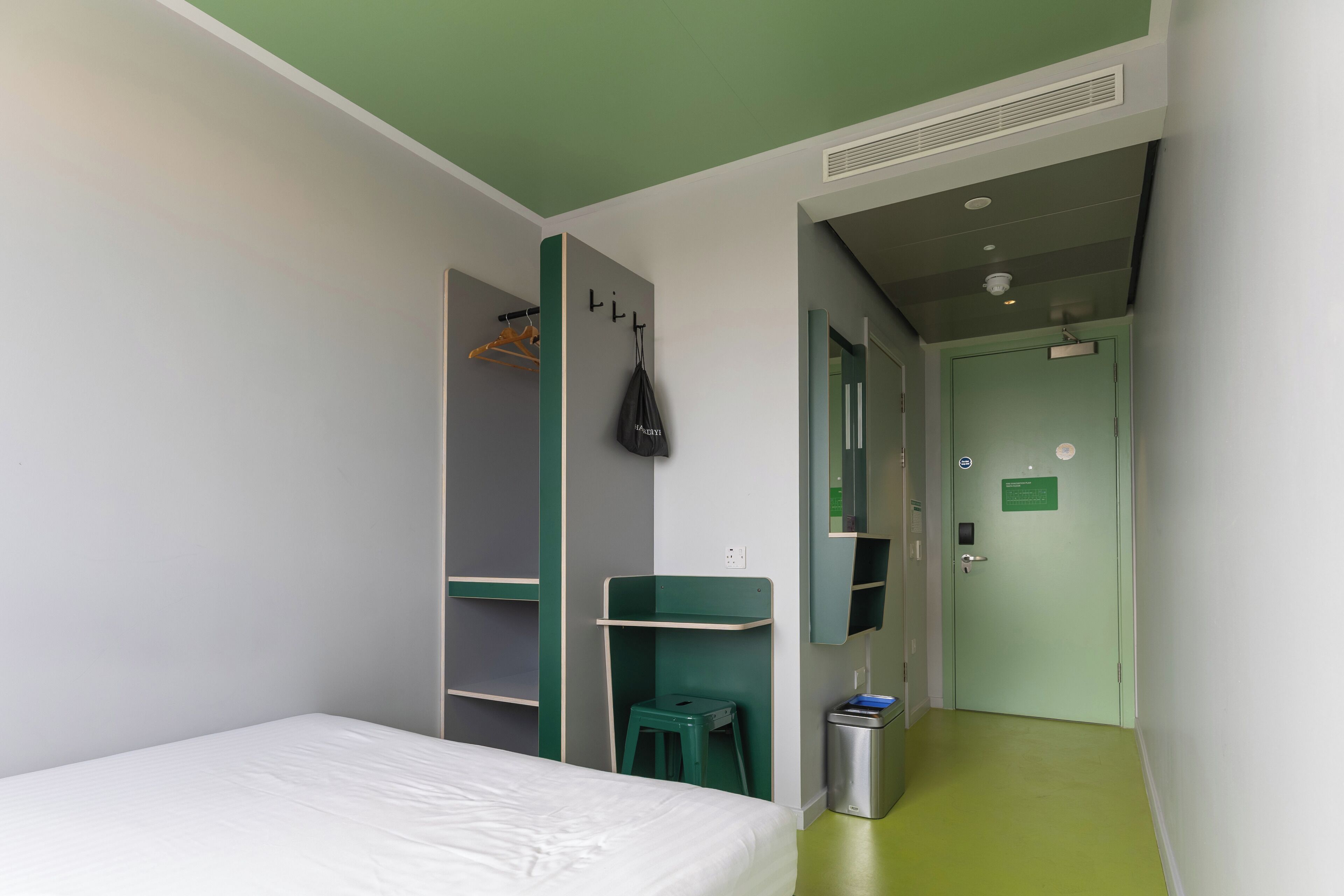 A room with a bed, a desk, a green stool, a trash bin, and a door with a sign.