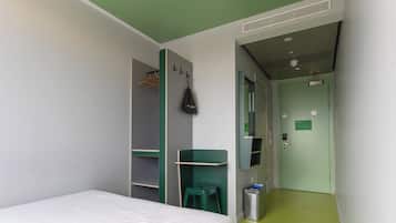 A room with a bed, a desk, a green stool, a trash bin, and a door with a sign.