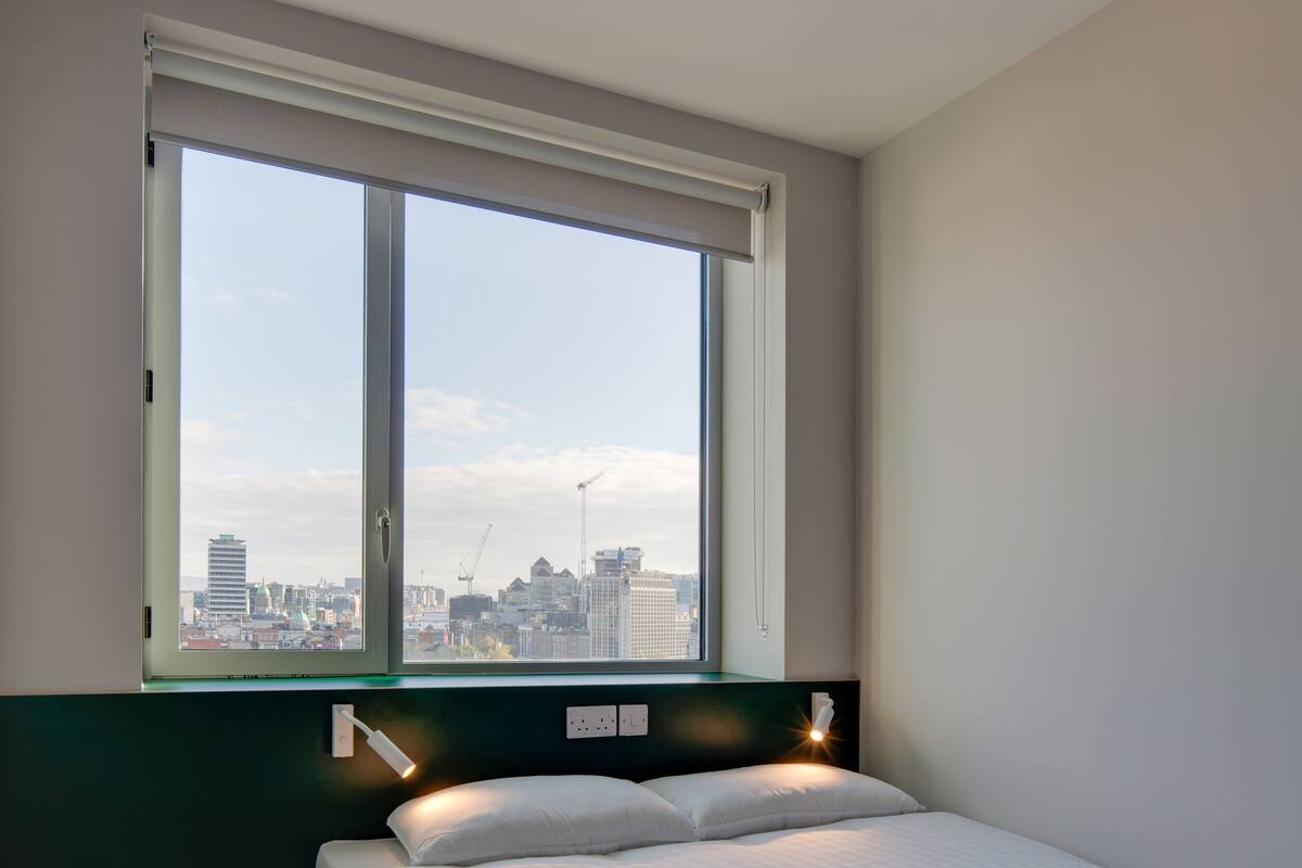 A modern hotel room with a large window, a neatly made bed, and a green floor.